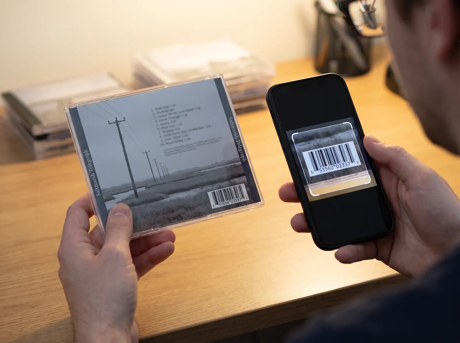 Illustration of a hand holding the back of a fictional CD, Saltmarsh Telegraph, with its tracklist, its title on both spines and a barcode in the corner, while a phone’s camera frames the barcode