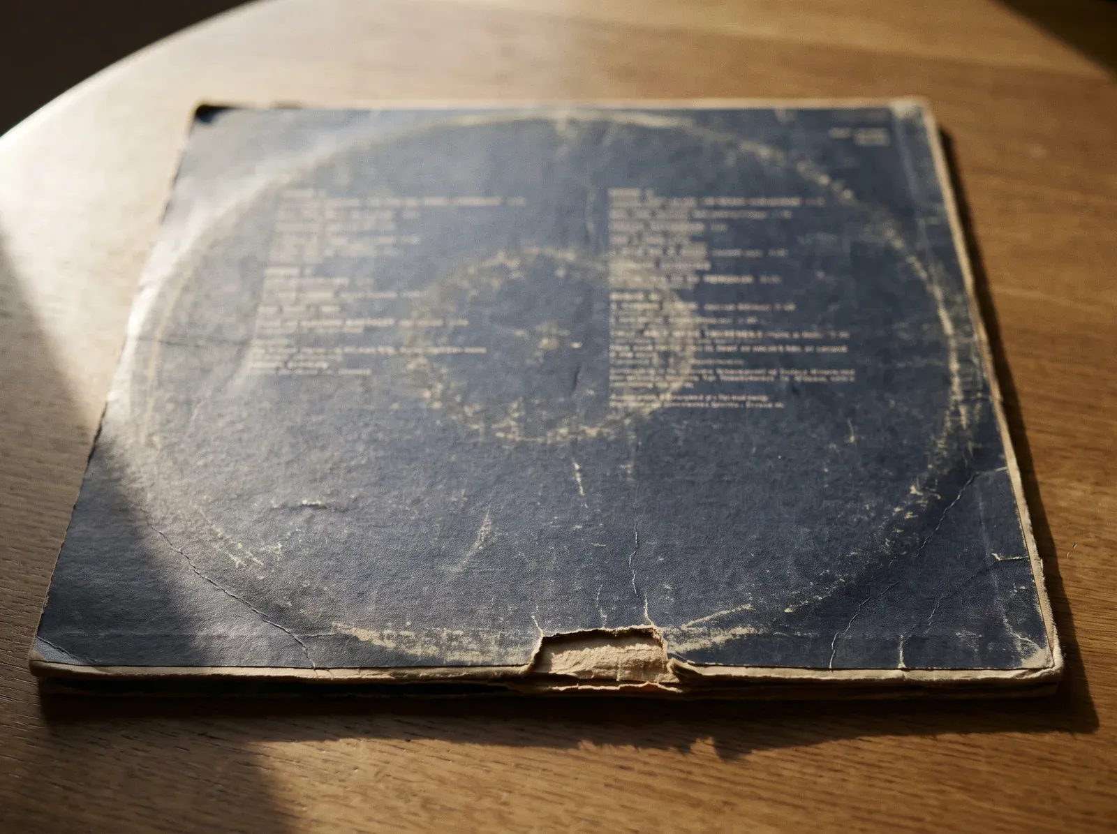 Illustration of the back of a worn LP sleeve: a pale ring of rubbed-off print where the record pressed against it, a long torn split in the middle of the bottom seam, fine creases and frayed edges and corners